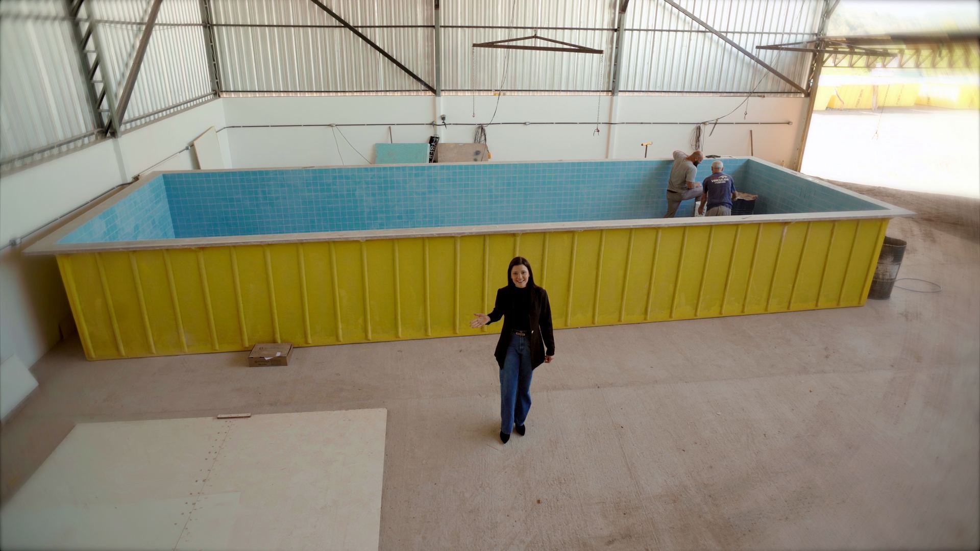 Apresentadora da Fyber Show em frente ao casco amarelo do Projeto Garden dentro da fábrica, com as paredes e o fundo já revestidos em pastilhas azuis e dois funcionários trabalhando na ponta